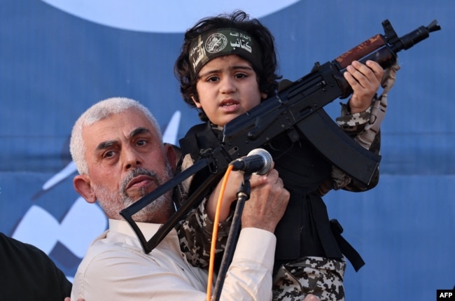 Pemimpin Hamas Yahya Sinwar menggendong anak seorang pejuang Brigade Al-Qassam, yang tewas dalam pertempuran baru-baru ini dengan Israel, dengan senapan Kalashnikov di tangannya selama unjuk rasa di Kota Gaza pada 24 Mei 2021. (Foto: voaindonesia.com/ AFP)